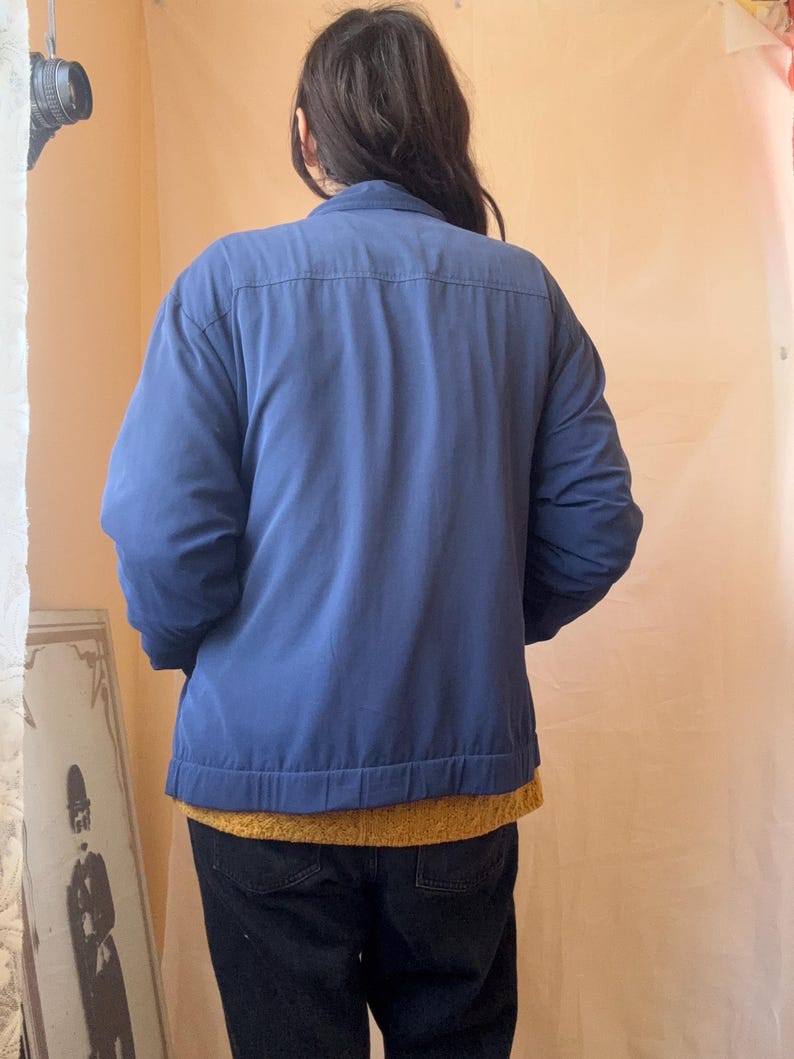 May include: A solid blue long-sleeved jacket with a collared neckline and a straight hemline. The jacket is worn over a mustard yellow textured sweater and dark-colored pants. The back view shows a horizontal seam across the upper back area.