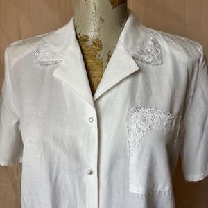 May include: A white short-sleeved button-down shirt featuring a pointed collar and a single chest pocket. The collar tips and the top corner of the chest pocket are adorned with white floral lace appliqués. The shirt has small, round, pearl-like buttons.