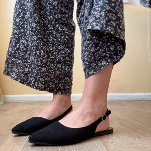 Könnte beinhalten: Ein Paar schwarze Slingback-Flats mit spitzer Zehenpartie und einer kleinen goldenen Schnalle. Die Schuhe sind aus einem weichen, strukturierten Material gefertigt. Die Schuhe werden auf einem hellen Holzboden präsentiert.