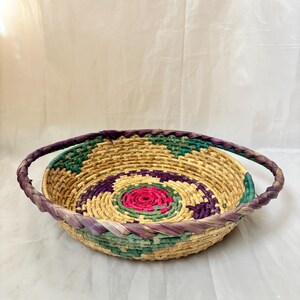 May include: A round, shallow woven basket featuring a coiled design with natural tan fibres accented by vibrant pink, purple, and teal geometric patterns. The basket includes two integrated braided handles in a deep purple hue along the rim.