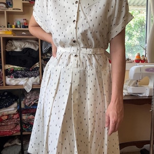 May include: Cream-colored dress with a black dotted pattern. The dress features short sleeves, a button-up front, and a belted waist. The pleated skirt falls to the mid-calf. The dress is made of a lightweight fabric, perfect for a vintage or retro look.