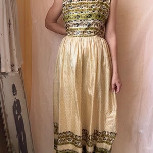 May include: A sleeveless floor-length dress featuring a square neckline and a fitted bodice. The bodice and hem are decorated with horizontal bands of green, black, and white geometric patterns. The skirt is a solid pale yellow with a subtle texture.