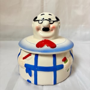 May include: A ceramic jar with a lid shaped like a bald man wearing black glasses and a mustache. The jar features a white base with blue grid lines, a red bowtie, and painted hands holding a red toothbrush and a red tube of toothpaste.