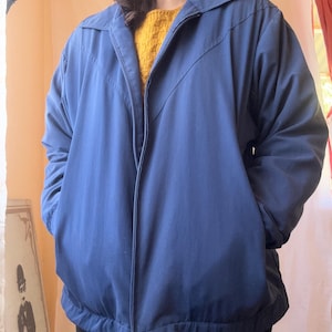 May include: A navy blue zip-up jacket featuring a pointed collar, long sleeves, and two front pockets. The jacket has a V-shaped seam detail across the chest and a ribbed hemline. A mustard yellow textured sweater is visible underneath the jacket.