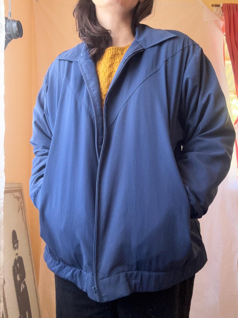 May include: A navy blue zip-up jacket featuring a pointed collar, long sleeves, and two front pockets. The jacket has a V-shaped seam detail across the chest and a ribbed hemline. A mustard yellow textured sweater is visible underneath the jacket.