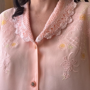 May include: A light peach-colored button-down blouse featuring a delicate lace collar with scalloped edges. The fabric is semi-sheer and decorated with embroidered floral patterns in shades of yellow and light pink, finished with round buttons.