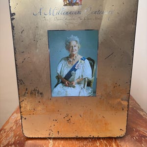 May include: A gold-coloured metal tin featuring a portrait of Queen Elizabeth II. The tin is inscribed with "A Millennium Centenary" and the years "1900-2000". The image of the Queen is set within a decorative frame.