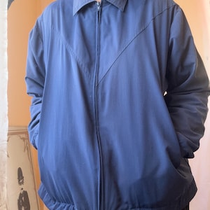 May include: A solid navy blue zip-up jacket featuring a classic collar, long sleeves, and a V-shaped seam detail across the chest. The jacket includes a full-length front zipper closure and an elasticized hemline for a structured fit.