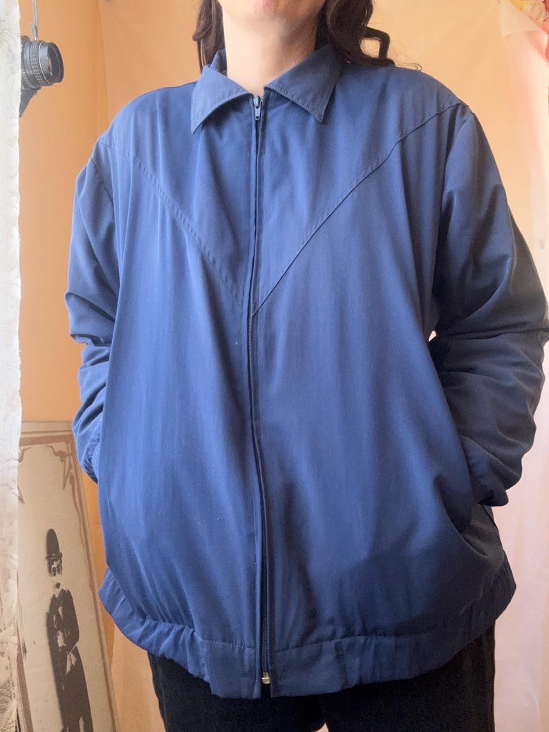 May include: A solid navy blue zip-up jacket featuring a classic collar, long sleeves, and a V-shaped seam detail across the chest. The jacket includes a full-length front zipper closure and an elasticized hemline for a structured fit.