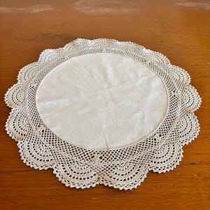 Könnte beinhalten: Ein rundes, weißes Stoffdeckchen mit einer glatten Mitte, die von einem aufwendigen, gehäkelten Spitzenrand mit wellenförmigem Abschluss umgeben ist. Die feine Handarbeit bildet ein wiederkehrendes Muster aus kleinen Löchern und Fächerformen.