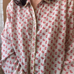 May include: A long-sleeved button-down shirt featuring a cream-colored base with a repeating pattern of small red paisley motifs and tiny black dots. The shirt includes a pointed collar and a vertical placket with light-colored buttons and yellow trim.