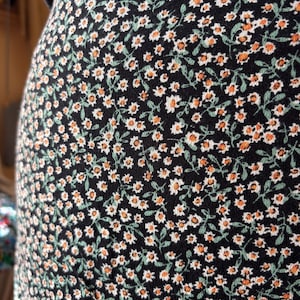 May include: Close-up of a black fabric covered in a repeating pattern of small white flowers with orange centers and green leaves. The floral design is set against a dark background. The fabric appears to be part of a garment.