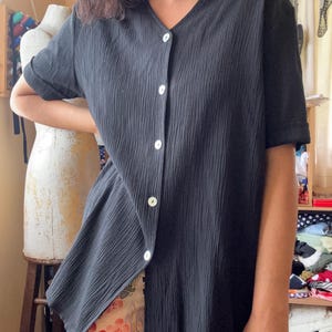May include: Black short-sleeved button-down shirt with a V-neck. The shirt has a textured, ribbed fabric and white buttons. The sleeves are cuffed. The shirt is paired with floral patterned pants. The shirt is a vintage style.
