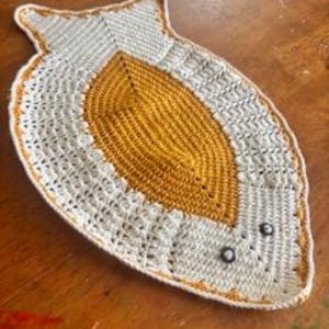 May include: A fish-shaped crochet doily featuring a central mustard yellow oval surrounded by off-white yarn. The design includes a textured stitch pattern, a mustard yellow border, and two small black beads serving as eyes near the head section.