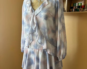 May include: A vintage-style dress on a mannequin. The dress features a V-neckline with ruffled trim, long sleeves, and a tiered skirt. The fabric has a watercolor-like pattern in shades of blue, pink, and white.