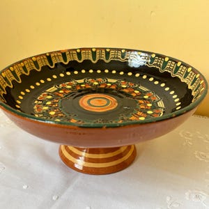 May include: A decorative, pedestal serving bowl with a wide, shallow dish and a short, round base. The bowl features a black interior with a colourful, ornate design in orange, yellow, and green. The exterior is a reddish-brown colour.