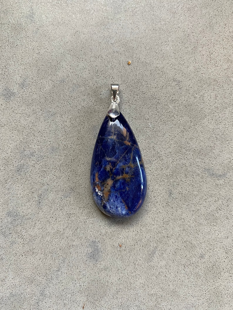 Puede incluir: Un colgante en forma de l&aacute;grima hecho de lapisl&aacute;zuli azul con motas doradas. El colgante est&aacute; unido a una anilla de plata.