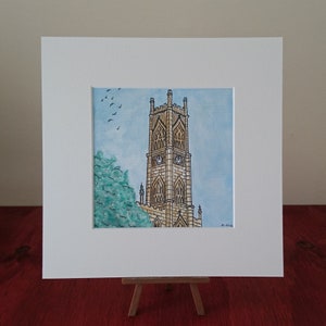Op de afbeelding: Een aquarel van een hoge, stenen kerktoren met een klok. De toren is afgebeeld tegen een lichtblauwe lucht met een paar vogels die erboven vliegen. Het schilderij is ingelijst in een witte passe-partout.