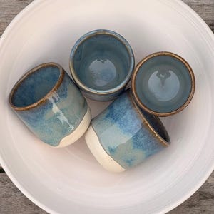 K&ouml;nnte beinhalten: Ein Set aus vier Keramikbechern mit blau-wei&szlig;er Glasur, die eine glatte, abgerundete Form und einen braunen Rand aufweisen. Die Becher sind in einer gro&szlig;en, schlichten wei&szlig;en Keramikschale angeordnet und zeigen ein gl&auml;nzendes Finish.