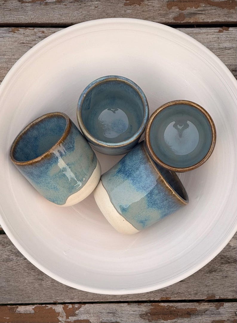K&ouml;nnte beinhalten: Ein Set aus vier Keramikbechern mit blau-wei&szlig;er Glasur, die eine glatte, abgerundete Form und einen braunen Rand aufweisen. Die Becher sind in einer gro&szlig;en, schlichten wei&szlig;en Keramikschale angeordnet und zeigen ein gl&auml;nzendes Finish.