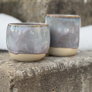 May include: Two ceramic cups with a gray and blue glaze and a tan base. The cups are different sizes, one short and wide, the other taller and narrower. The cups are sitting on a weathered stone surface.