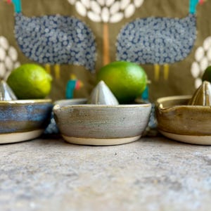 May include: Three ceramic lime juicers in different colors: blue, gray, and brown. Each juicer has a white rim and a textured surface. The juicers are arranged in a row on a gray surface.