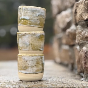 K&ouml;nnte beinhalten: Ein Stapel aus drei zylindrischen Keramikbechern mit einer gesprenkelten Glasur in Olivgr&uuml;n, Wei&szlig; und Beige. Die Becher haben einen glatten, unglasierten Boden und eine texturierte, gesprenkelte Oberfl&auml;che im oberen Bereich der Gef&auml;&szlig;e.