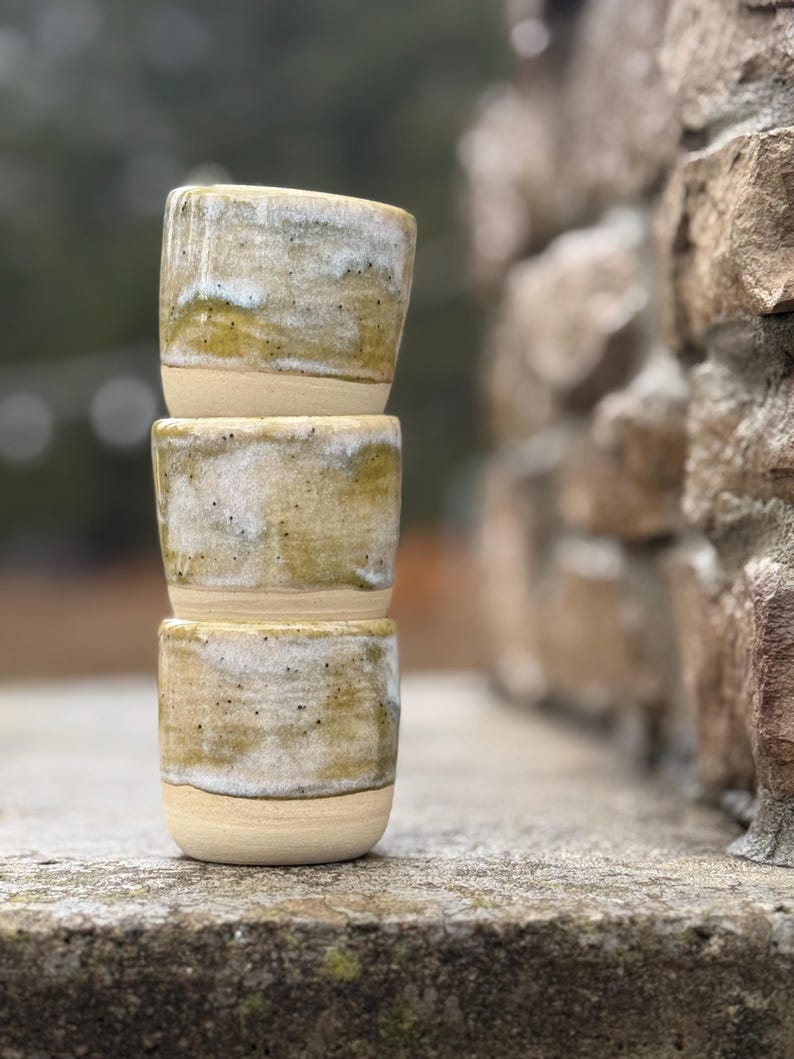 K&ouml;nnte beinhalten: Ein Stapel aus drei zylindrischen Keramikbechern mit einer gesprenkelten Glasur in Olivgr&uuml;n, Wei&szlig; und Beige. Die Becher haben einen glatten, unglasierten Boden und eine texturierte, gesprenkelte Oberfl&auml;che im oberen Bereich der Gef&auml;&szlig;e.