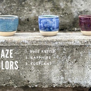 K&ouml;nnte beinhalten: Drei kleine Keramikbecher mit einer glatten, unglasierten beigen Basis und gl&auml;nzenden oberen Abschnitten in den Farben Blaurutil, Saphir und Aubergine. Der Text GLAZE COLORS steht links, mit den Nummern 1, 2 und 3 f&uuml;r die jeweiligen Farben.
