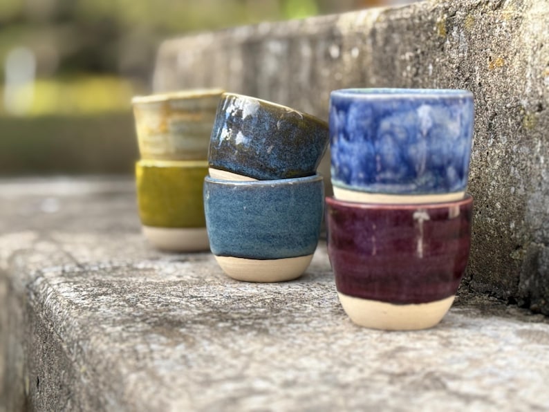 K&ouml;nnte beinhalten: Eine Sammlung von f&uuml;nf kleinen Keramikbechern mit glasierten Oberfl&auml;chen in Blau-, Lila-, Gelb- und Gr&uuml;nt&ouml;nen. Jeder Becher hat einen glatten, unglasierten beigen Boden und ein abgerundetes, henkelloses Design, angeordnet in zwei Stapeln.