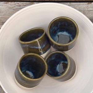 K&ouml;nnte beinhalten: Vier Keramikbecher in einer Gruppe angeordnet, jeder mit einer gl&auml;nzenden Oberfl&auml;che mit einem Farbverlauf in tiefem Olivgr&uuml;n und Dunkelblau im oberen Bereich und einer einfarbigen cremewei&szlig;en Basis. Die Becher haben eine zylindrische Form.