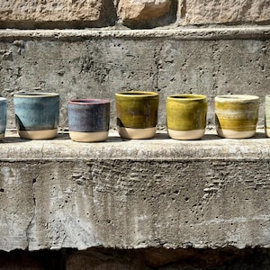 May include: A row of seven ceramic cups with a cylindrical shape, each featuring a smooth, unglazed beige base and a glossy, multi-colored glazed upper section in shades of blue, purple, olive green, and pale yellow, arranged in a line.