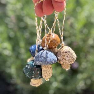 Könnte beinhalten: Eine Sammlung von Keramik-Pilzornamenten in verschiedenen Farben, darunter Blau, Braun und Grün, jeweils mit einem einzigartigen Sprenkelmuster. Die Ornamente sind an weiß-goldenen Schnüren aufgehängt, ideal für die Feiertags- oder Saison-Dekoration.