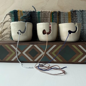 May include: Three white ceramic yarn bowls with different colored yarn coming out of them. The bowls are sitting on a wooden shelf with a geometric pattern.