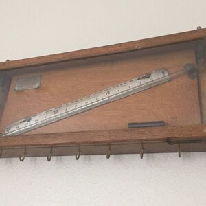 Può includere: Un dispositivo vintage in legno incorniciato con un fronte in vetro, che presenta un righello in metallo e un tubo di vetro con un'asta di metallo all'interno. Il dispositivo è montato su una parete con ganci e una manovella in metallo.