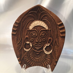 May include: A brown, carved wooden decorative plate featuring a stylized face with large hoop earrings and a headband. The face has detailed features and is set against a textured background. The plate is displayed on a clear stand.