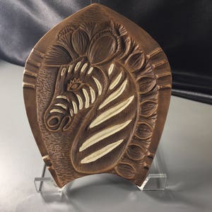 May include: A brown, carved wooden plaque featuring a zebra head with cream-colored stripes. The plaque has a decorative border and is mounted on a clear stand. The zebra's eye is detailed, and the overall design is artistic.