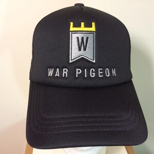 May include: Black trucker hat with a flat brim. The hat features an embroidered design of a yellow crown above a silver banner with a white "W" and the text "WAR PIGEON" in silver.