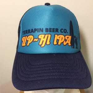 May include: A blue and navy trucker hat with the text "TERRAPIN BEER CO. UP~HI IPA" in orange and white. The hat features a black mesh back and a curved brim. A silhouette of a surfboard and person is on the right side of the hat.
