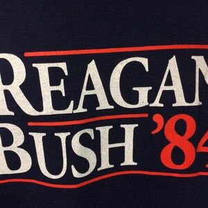 May include: A navy blue t-shirt with white text that reads "Reagan Bush '84" in a red, white, and blue color scheme.