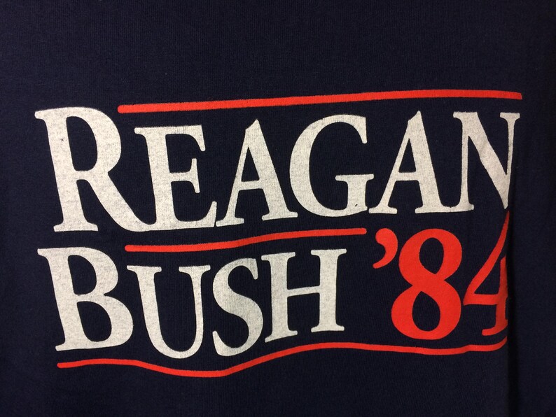 May include: A navy blue t-shirt with white text that reads "Reagan Bush '84" in a red, white, and blue color scheme.