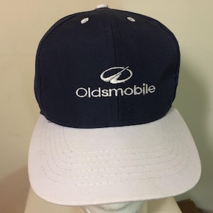May include: A blue and white baseball cap with the Oldsmobile logo and the word "Oldsmobile" embroidered in white on the front.