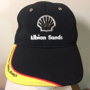 May include: Black baseball cap with a yellow and red brim. The cap has a silver Shell logo and the text "Albion Sands" embroidered on the front. The brim has the text "Always Home Safely, Always" embroidered on it.