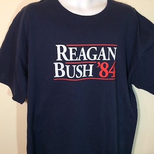 May include: A navy blue t-shirt with the text "Reagan Bush '84" printed in white and red.