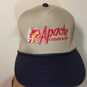 May include: A tan and navy blue baseball cap with an embroidered logo that says "Apache Construction" and a red, white, and yellow image of an American Indian chief's head.
