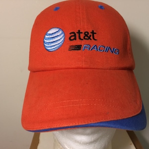 May include: Orange baseball cap with a blue brim and accents. The cap features the AT&T logo and the word "RACING" embroidered in blue. The cap is made of a durable fabric.