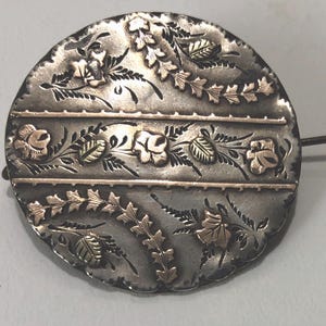 May include: A round, antique brooch with a silver-tone base and gold-tone floral accents. The brooch features a detailed design of roses and leaves, with horizontal bands. A pin extends from the back.