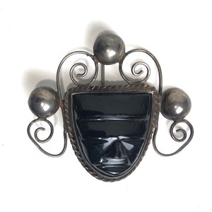 May include: Vintage silver-tone brooch featuring a carved black stone face within a decorative frame. The brooch has three silver spheres and ornate scrollwork. The face has carved details and a textured border.