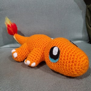 May include: An orange crochet Charmander plush toy with white accents on its feet and a flame tail. The toy has large black and white eyes with blue highlights. The plush is lying on a gray surface.