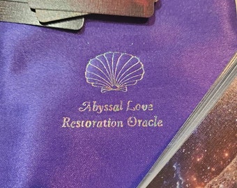 May include: A purple satin drawstring pouch featuring a gold foil seashell icon and the text Abyssal Love Restoration Oracle. A stack of oracle cards with space-themed imagery and text is partially visible on top of the pouch and to the side.